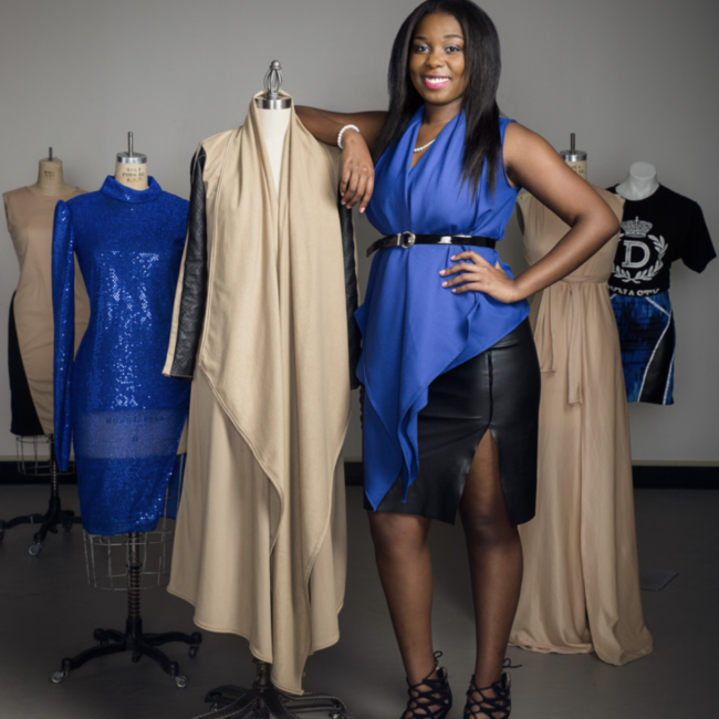 Britany in a royal blue shawl and leather skirt standing with her arm on the shoulder a mannequin wearing her design, 4 more mannequins are behind in a triangle formation